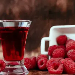 Szklaneczka z rubinową nalewką z malin, obok świeże maliny wysypujące się z białego naczynia.