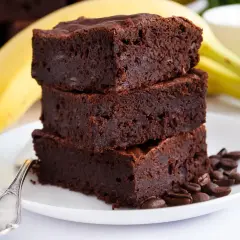 Stos pysznych brownie z bananami, ozdobiony ziarnami kawy, na talerzu z widocznym bananem w tle.