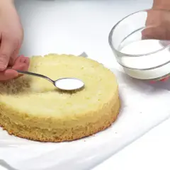 Ręce dodają łyżeczką biały płyn na biszkopt. To poncz do tortu, który sprawi, że będzie wilgotny i pyszny.