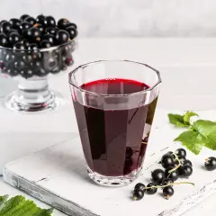 Szklanka z nalewką z czarnej porzeczki, obok świeże owoce i liście. W tle wazka pełna czarnych porzeczek.