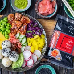 Kolorowy bowl z kawałkami łososia, edamame, mango, awokado, ryżowymi kulkami i marynowanym imbirem. Obok opakowanie różowego imbiru.