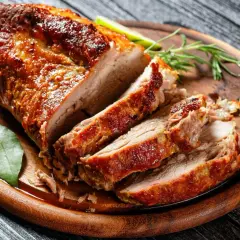 Soczysty schab pieczony na desce, pokrojony w plastry, z ziołami i liściem laurowym. Idealny przykład, jak upiec schab, żeby był soczysty.