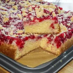Puszyste ciasto drożdżowe z malinami i kruszonką, jeden kawałek podniesiony, by pokazać jego strukturę.