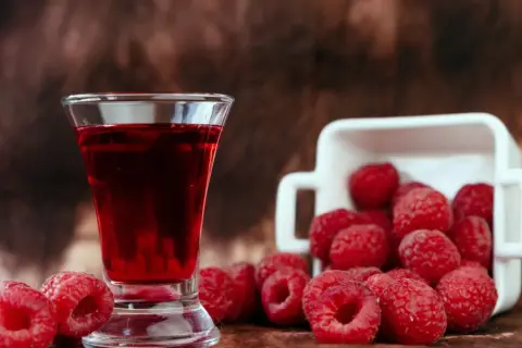 Szklaneczka z rubinową nalewką z malin, obok świeże maliny wysypujące się z białego naczynia.