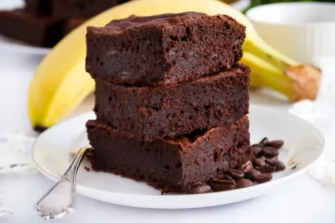 Stos pysznych brownie z bananami, ozdobiony ziarnami kawy, na talerzu z widocznym bananem w tle.
