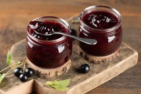 Dwa słoiczki domowego dżemu z czarnej porzeczki, jeden z łyżeczką pełną gęstego, ciemnego dżemu. Obok świeże owoce i listki.