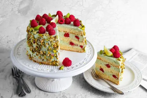 Pyszny tort pistacjowo malinowy, udekorowany świeżymi malinami i pistacjami, kusi swoim wyglądem i smakiem.