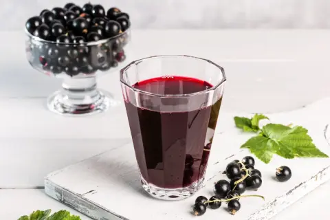 Szklanka z nalewką z czarnej porzeczki, obok świeże owoce i liście. W tle wazka pełna czarnych porzeczek.