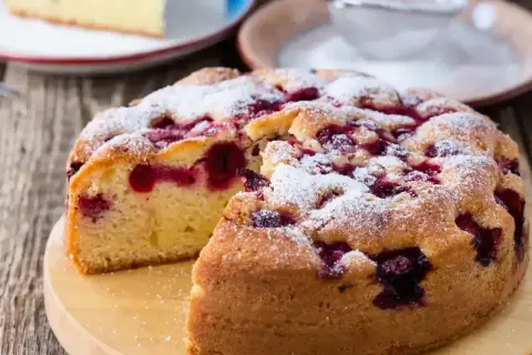 Wilgotne ciasto z wiśniami, posypane cukrem pudrem, wygląda apetycznie. Kawałek ciasta na talerzyku obok.