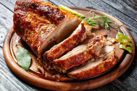 Soczysty schab pieczony na desce, pokrojony w plastry, z ziołami i liściem laurowym. Idealny przykład, jak upiec schab, żeby był soczysty.