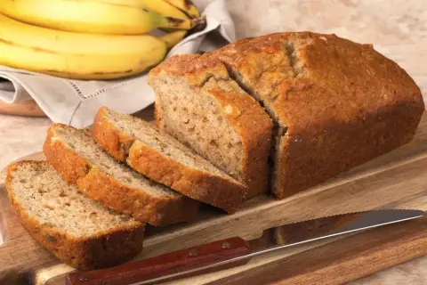 Pyszny chlebek bananowy bez cukru, pokrojony na kromki, leży na desce obok pęczka bananów.