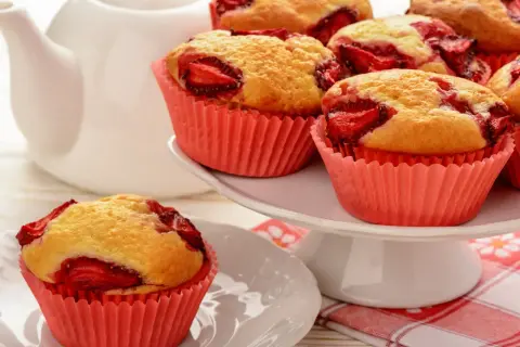 Pyszne muffinki z truskawkami w różowych papilotkach, obok białego dzbanka. Idealne na deser.