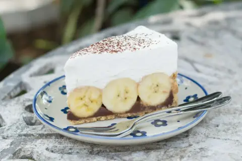 Pyszne ciasto bananowe bez pieczenia z kremową pianką i bananami na wierzchu, posypane kakao. Idealne na deser.