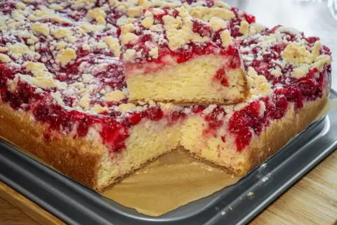Puszyste ciasto drożdżowe z malinami i kruszonką, jeden kawałek podniesiony, by pokazać jego strukturę.