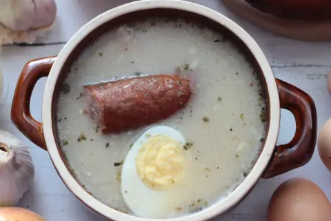 Aromaticzny żurek z kiełbasą i jajkiem, przygotowany na bazie domowego zakwasu na żurek. Obok czosnek, cebula i jajka.