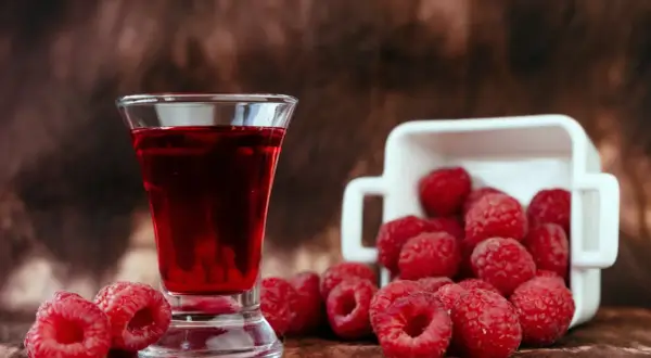Szklaneczka z rubinową nalewką z malin, obok świeże maliny wysypujące się z białego naczynia.