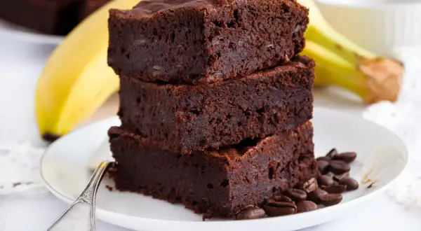 Stos pysznych brownie z bananami, ozdobiony ziarnami kawy, na talerzu z widocznym bananem w tle.