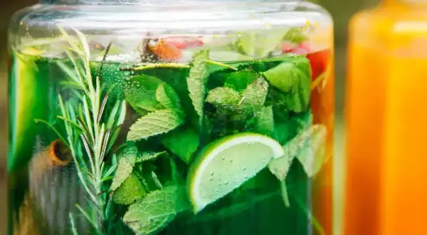 Zielony, orzeźwiający syrop z mięty w szklanym słoju, z dodatkiem limonki i rozmarynu. Obok pomarańczowy napój.