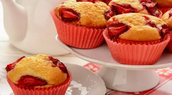 Pyszne muffinki z truskawkami w różowych papilotkach, obok białego dzbanka. Idealne na deser.