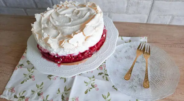 Pyszna tarta z porzeczkami, udekorowana puszystą bezą. Obok talerzyk z dwoma złotymi widelczykami.