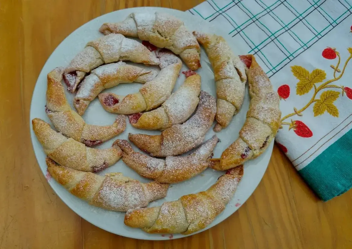 Pyszne, domowe rogaliki z marmoladą, posypane cukrem pudrem, ułożone na talerzu obok ręcznika z truskawkami.