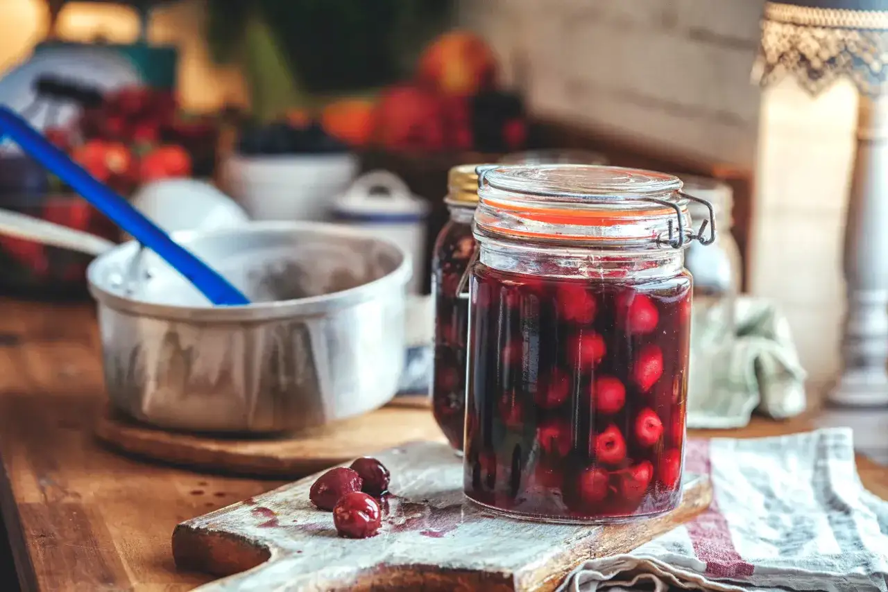 Słoik pełen domowego kompotu z wiśni, obok garnek i kilka owoców. Letnie przetwory gotowe do spiżarni.