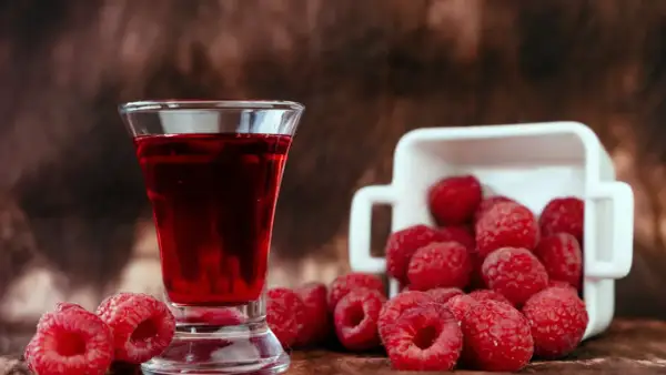 Szklaneczka z rubinową nalewką z malin, obok świeże maliny wysypujące się z białego naczynia.
