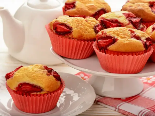 Pyszne muffinki z truskawkami w różowych papilotkach, obok białego dzbanka. Idealne na deser.