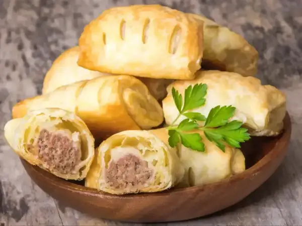 Stos pieczonego ciasta francuskiego z kiełbasą, ozdobiony natką pietruszki. Idealne na szybką przekąskę.