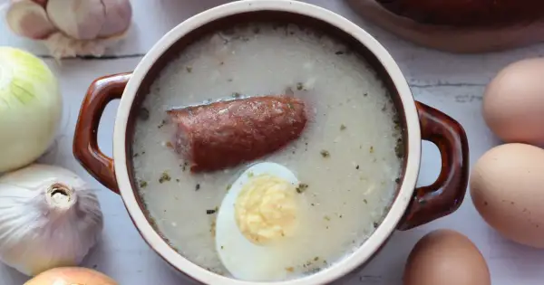 Aromaticzny żurek z kiełbasą i jajkiem, przygotowany na bazie domowego zakwasu na żurek. Obok czosnek, cebula i jajka.
